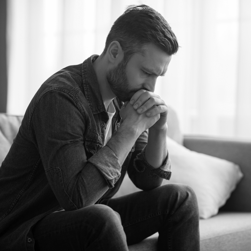 B&W image of a caucasian man sitting on the couch with his hand clasped to his chin